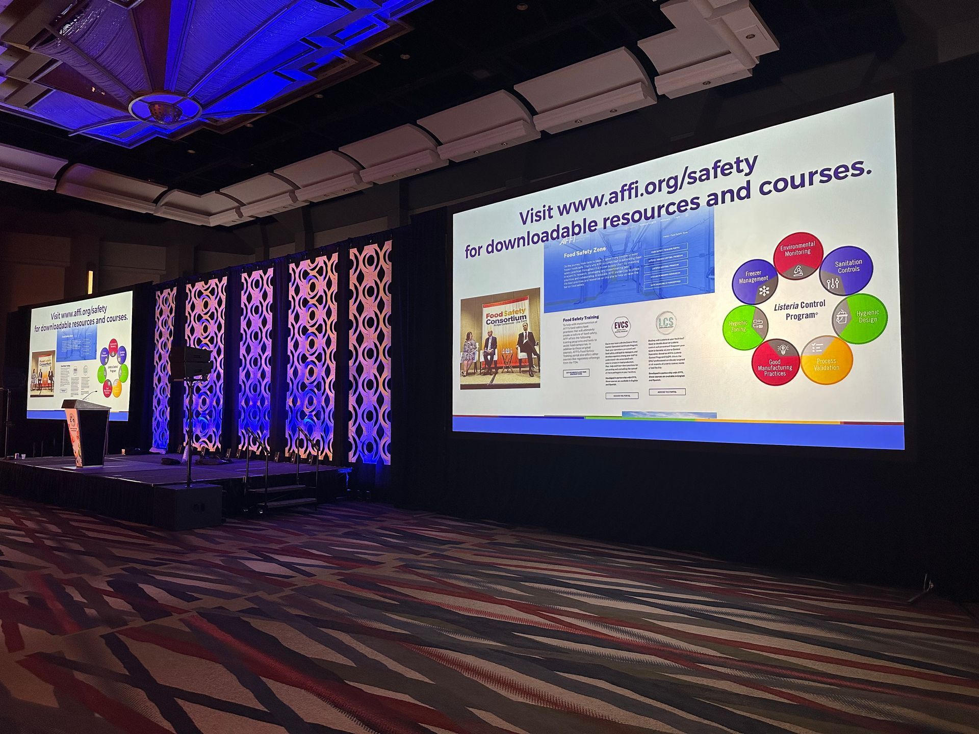 Conference hall with two large presentation screens showing a colorful slide and patterned carpeted floor