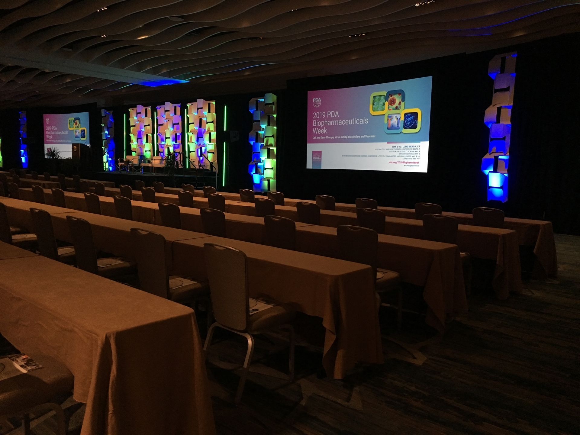 Conference hall with rows of tables and chairs, colorful stage lights, and presentation screens at the front.