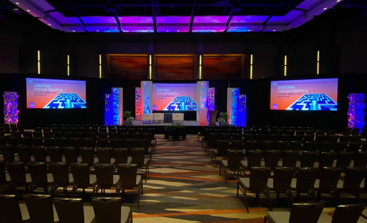 Empty conference hall with rows of chairs and three large screens on a stage lit in purple and orange.