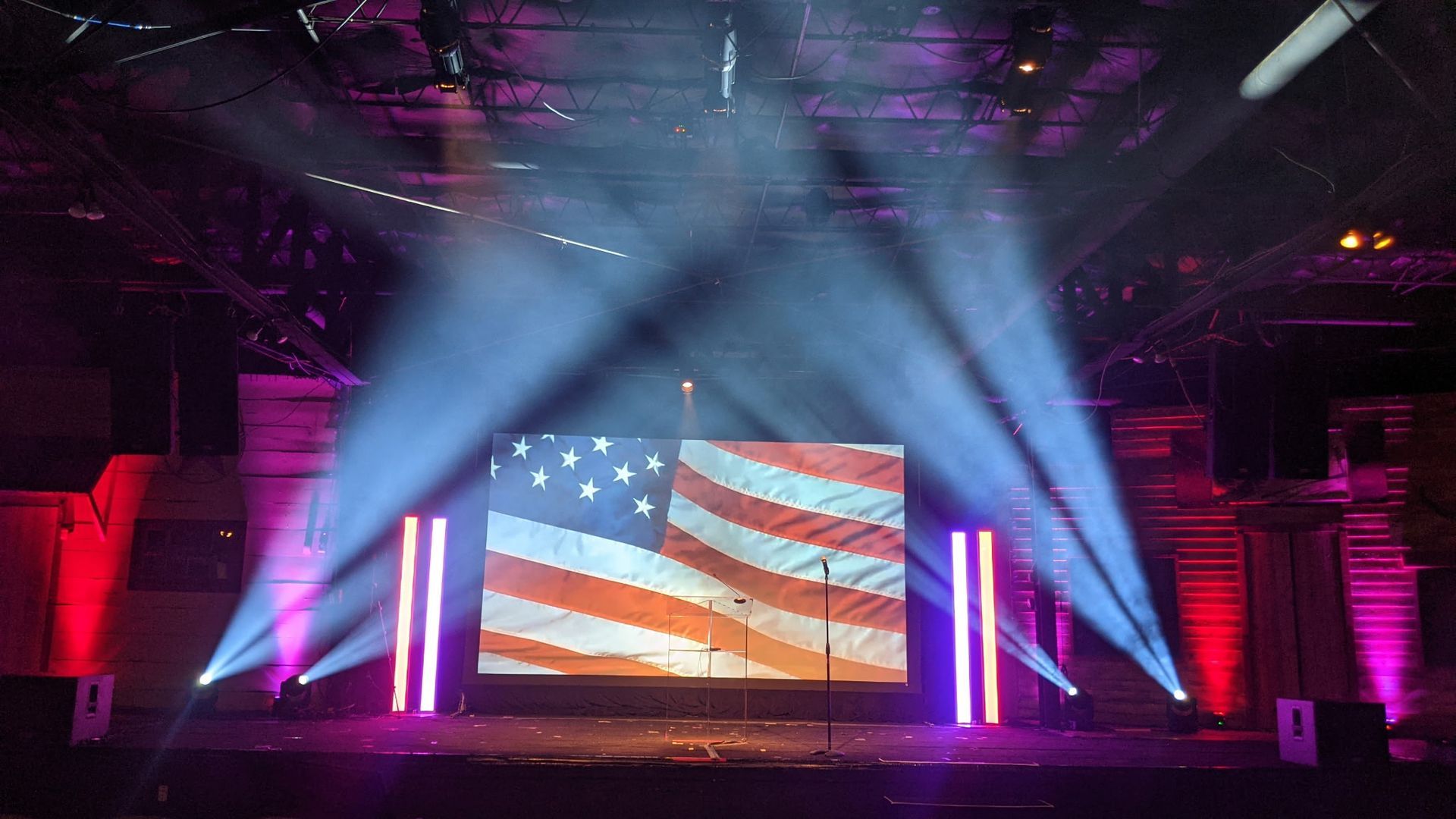 Concert stage with American flag on screen, purple lighting, and bright spotlights in a dark venue