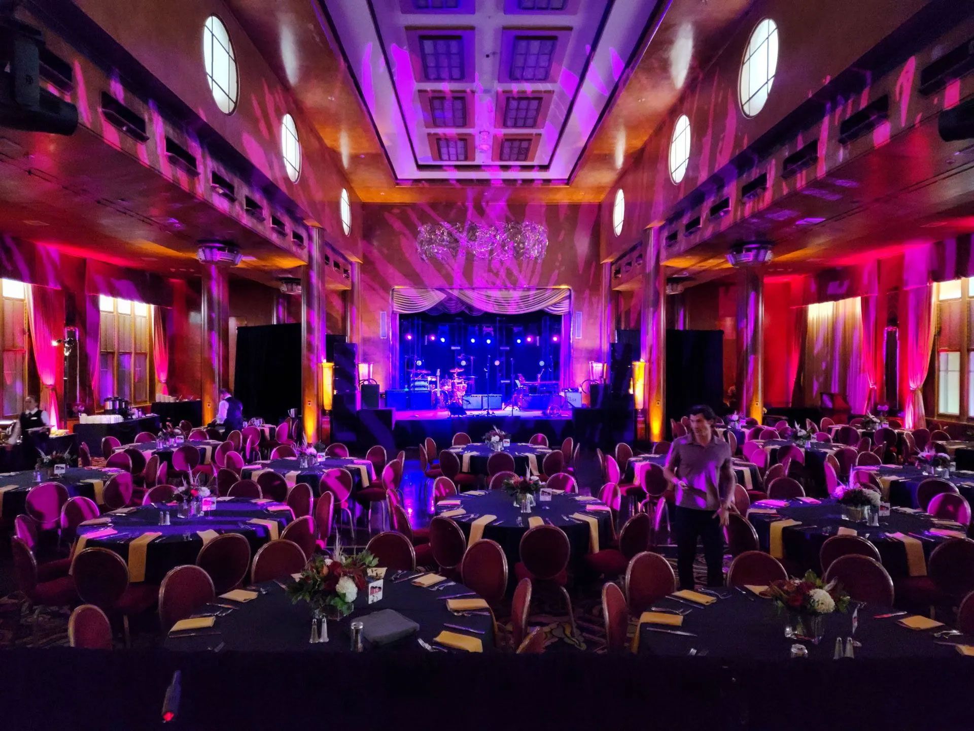 Crowded banquet hall lit in purple and pink, with guests facing a brightly lit stage at the far end