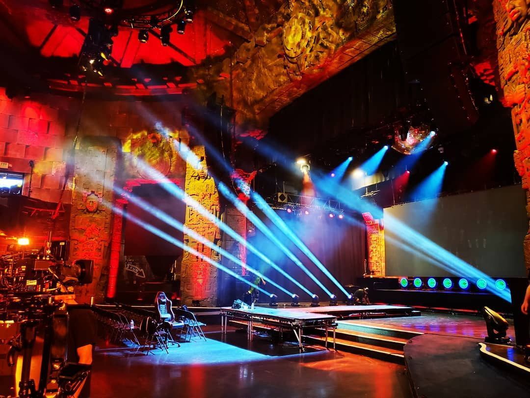 A brightly lit stage with red textured walls, illuminated by rows of sharp blue spotlights angled toward the floor.