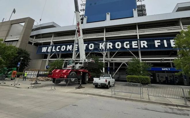 Street view of Kroger Field entrance with large welcome sign, trucks, and parked cars beside the stadium.