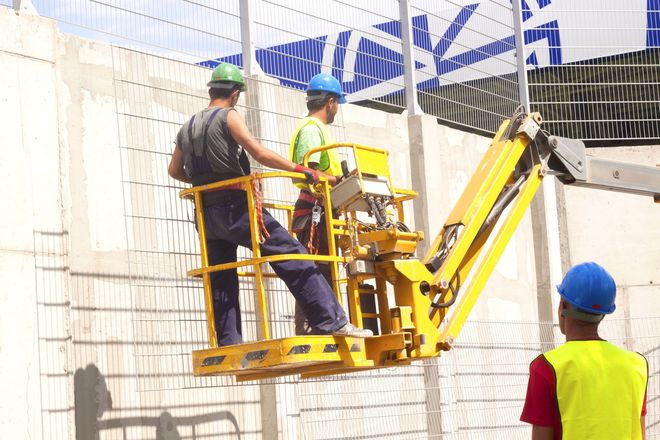 Workers operating a yellow aerial lift against a white tiled wall, with a worker in a blue hard hat nearby