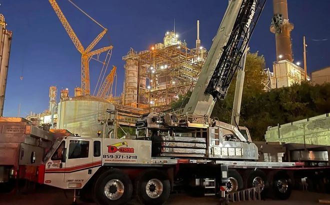 Crane truck at night near a lit construction site with tall cranes and industrial equipment.