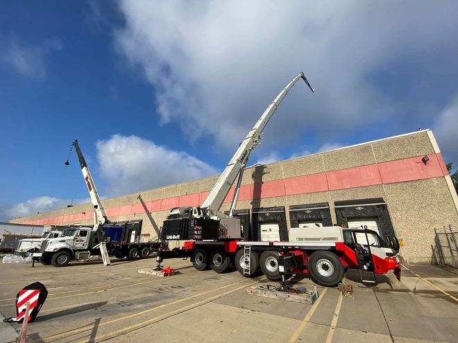 Two cranes lifting equipment beside a brick building under a blue sky
