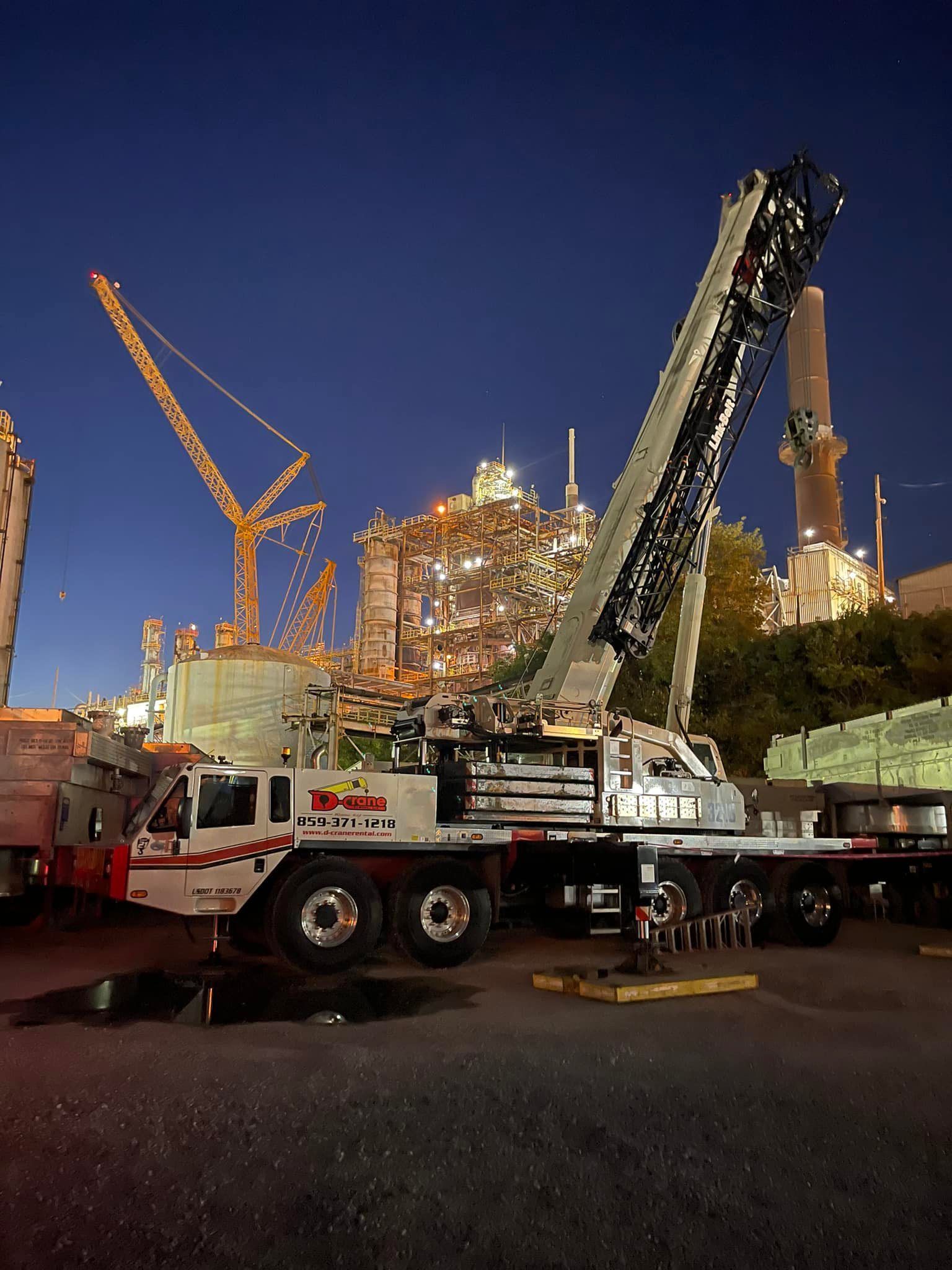 Crane lifting equipment at a construction site at dusk, with trucks and a lit building in the background