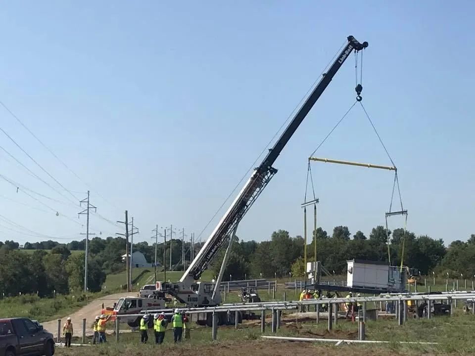 Crane lifting a large metal frame at a roadside worksite with workers in safety vests nearby