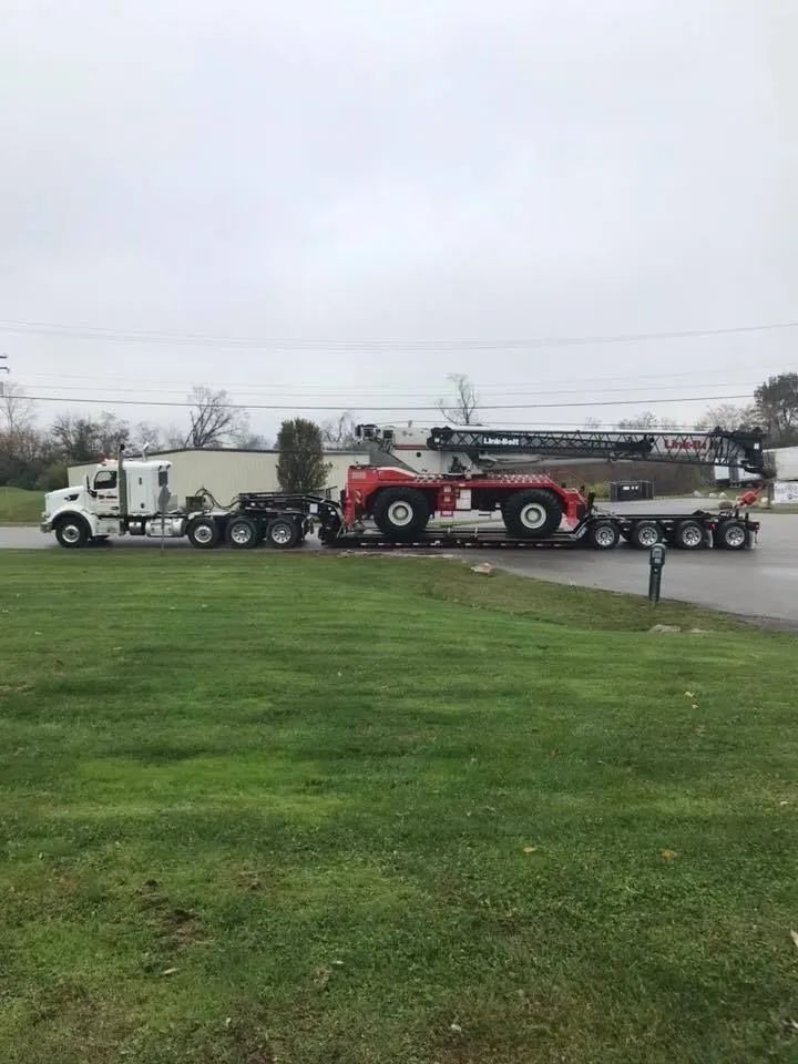 Red tractor on a flatbed trailer being towed by a white semi-truck in a grassy lot