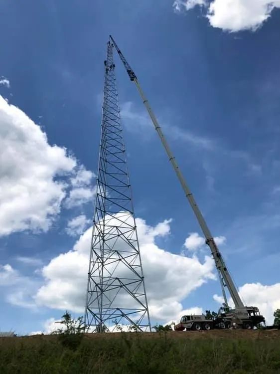Tall steel lattice tower being lifted by a crane under a blue sky with clouds.