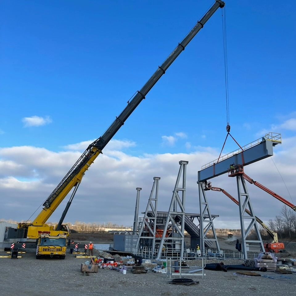 Two cranes lifting a large steel structure at a waterfront construction site under a blue sky