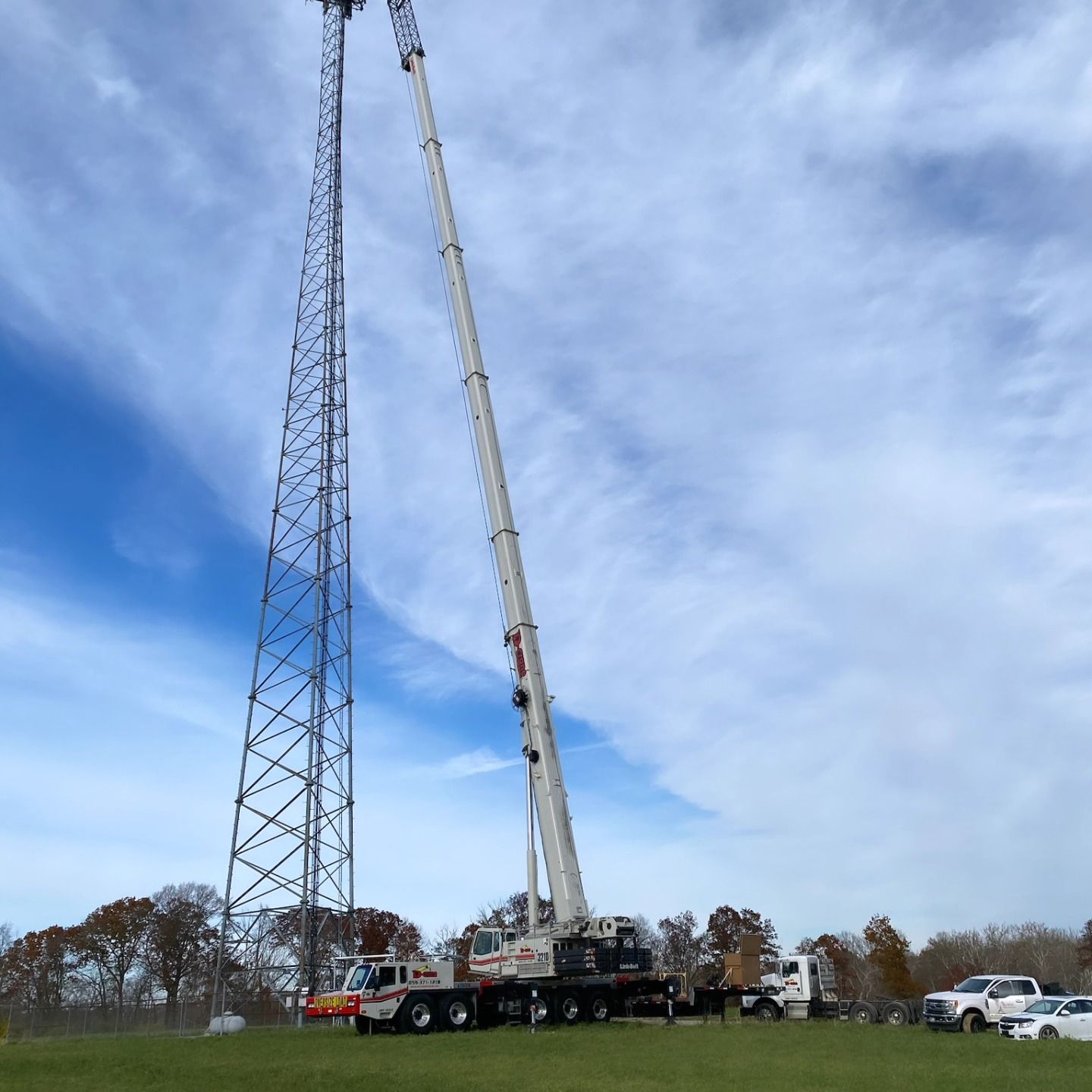 Crane lifting a tall radio tower section beside an existing lattice tower in a grassy field