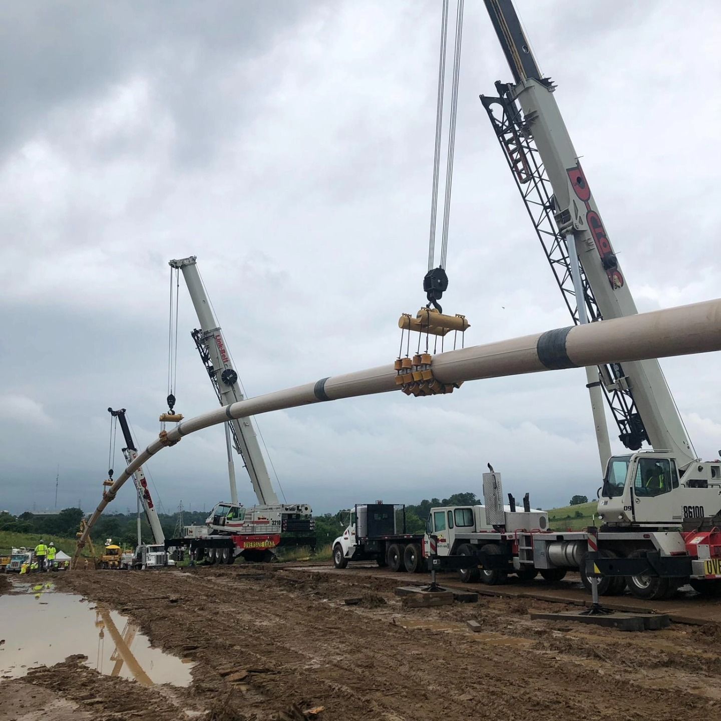 Cranes lifting a long white pipe at a muddy construction site under cloudy skies