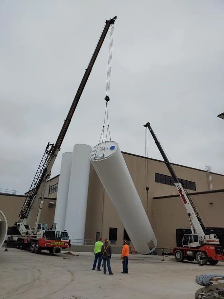 Crane lowering a large white cylindrical tank into place outside a building, with workers nearby.