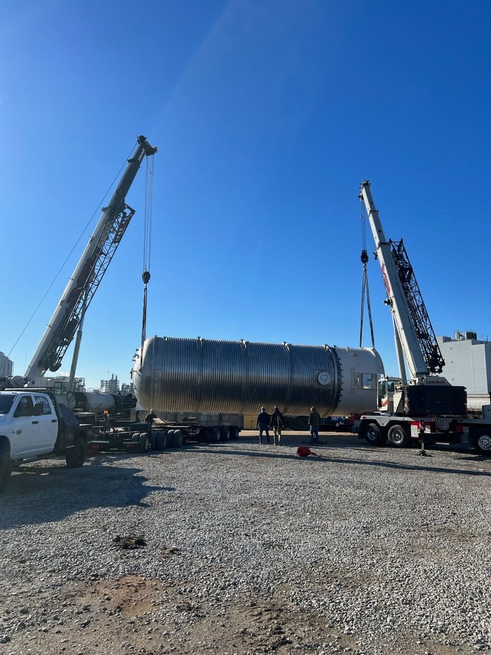 Two cranes lifting a large metal tank at a gravel construction site under a blue sky