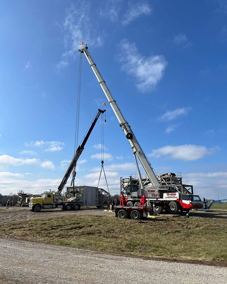 Two cranes lifting in a grassy field under a blue sky, with a truck nearby.
