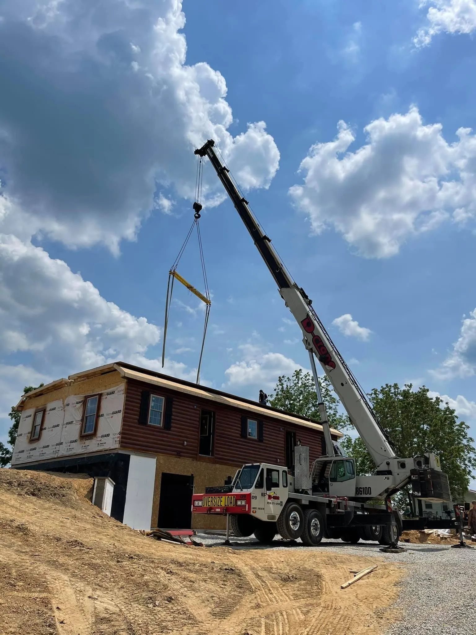 Crane lifting a two-story house frame at a construction site under a cloudy blue sky