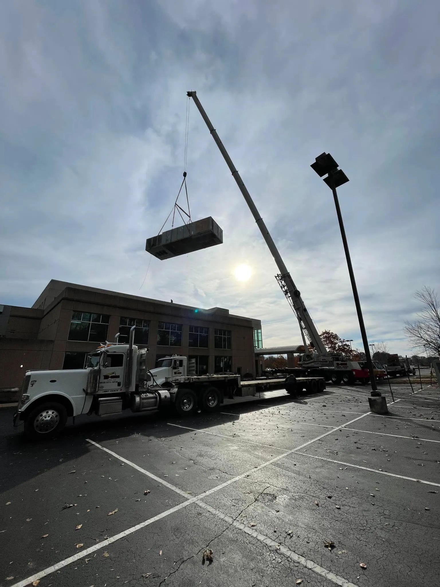 Crane lifting a large HVAC unit beside a building in a wet parking lot under cloudy skies