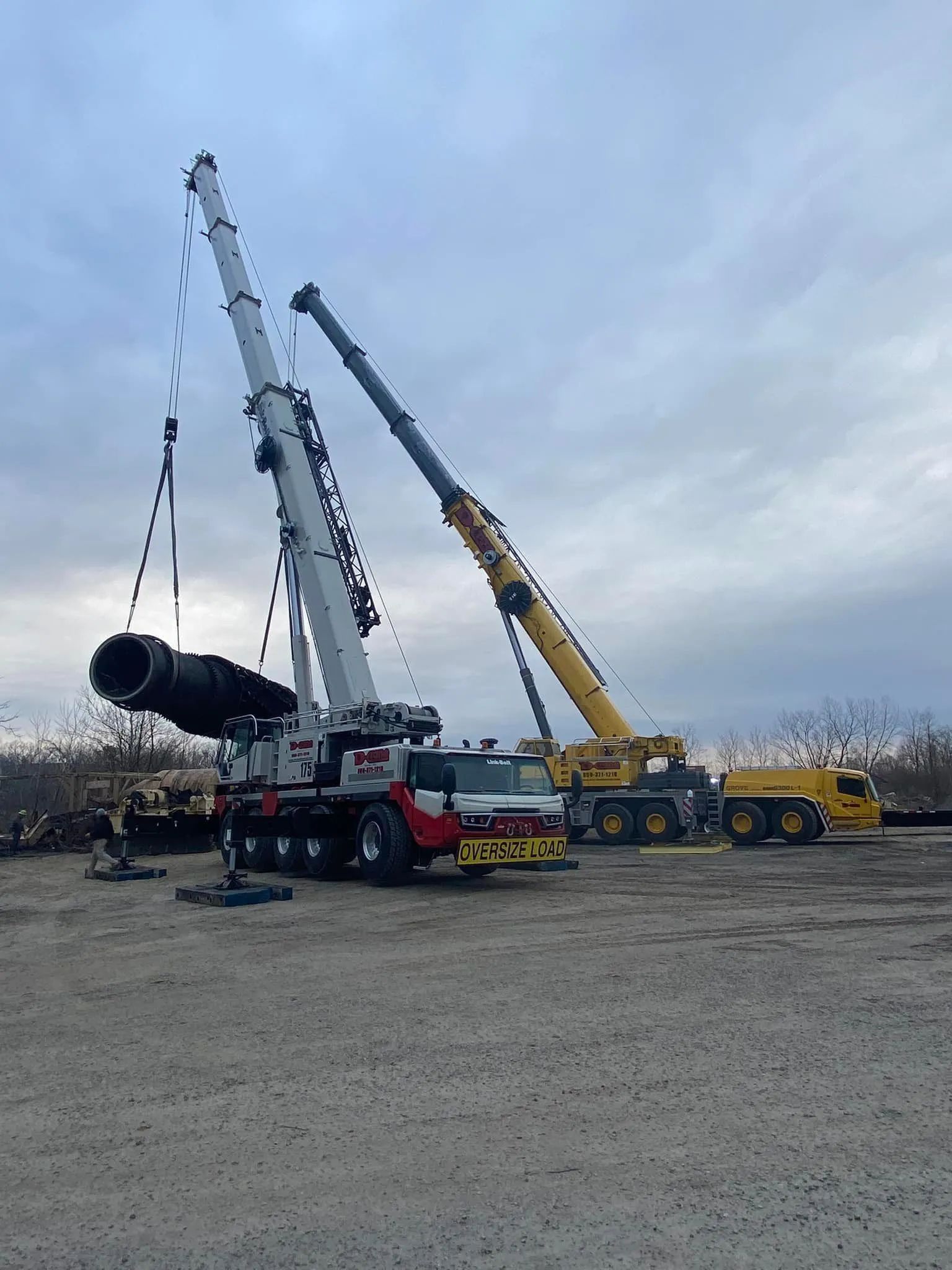 Three cranes lifting a large black pipe in a gravel lot under a cloudy sky