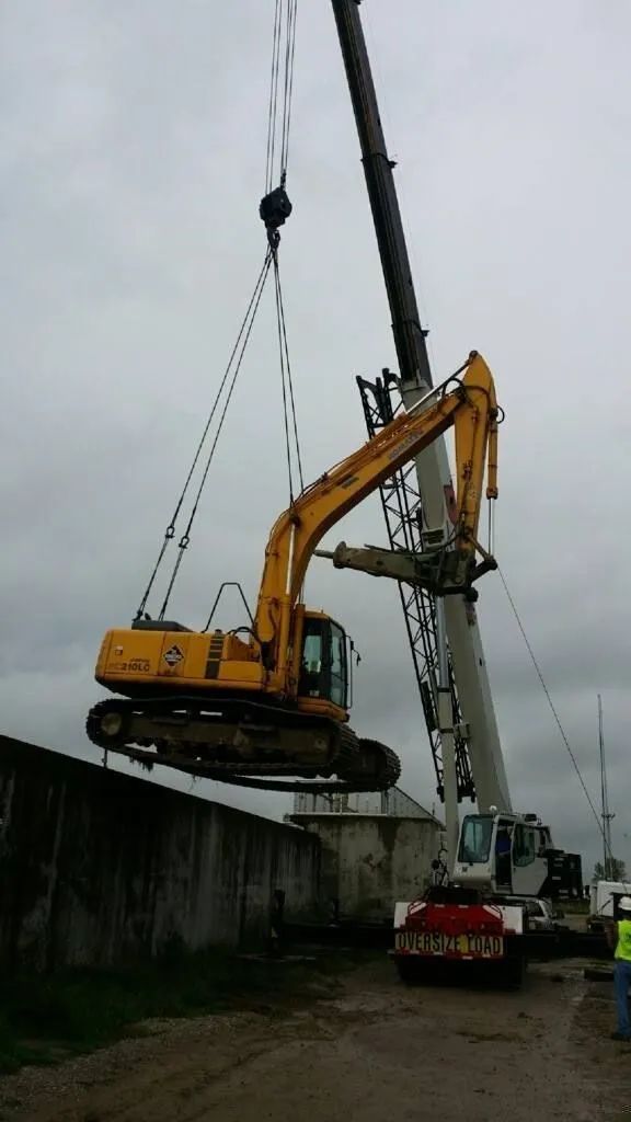 Yellow excavator being lifted by a crane beside a concrete wall on an overcast construction site