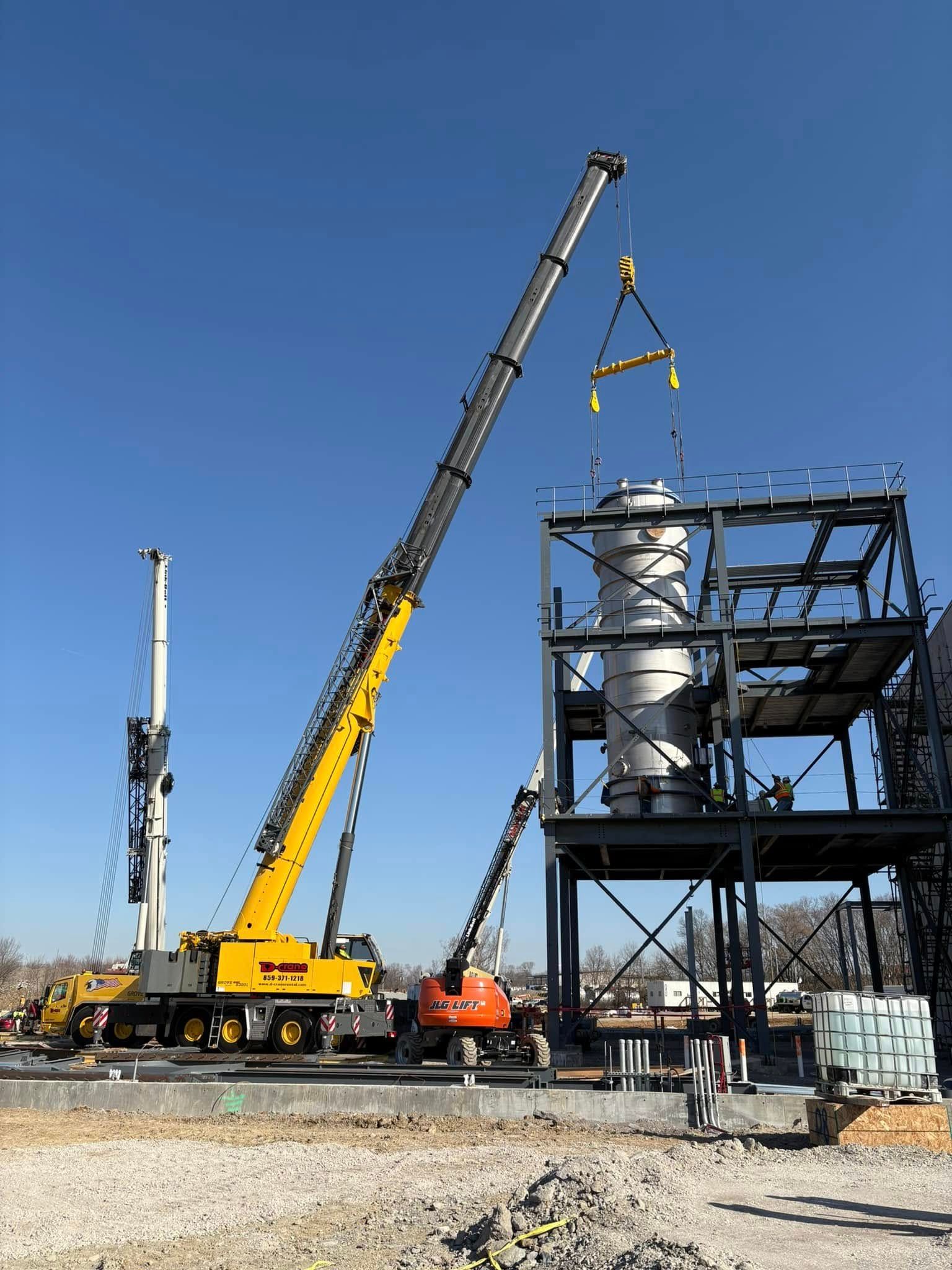 Crane lifting a large silver tank beside a steel industrial structure under a clear blue sky