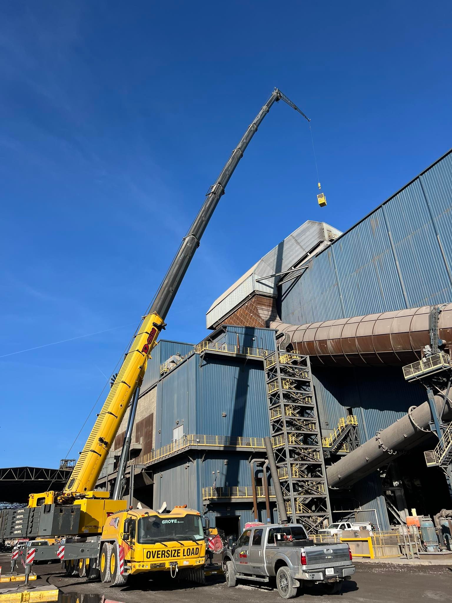 Crane lifting materials beside a large blue stadium under construction, with trucks below.
