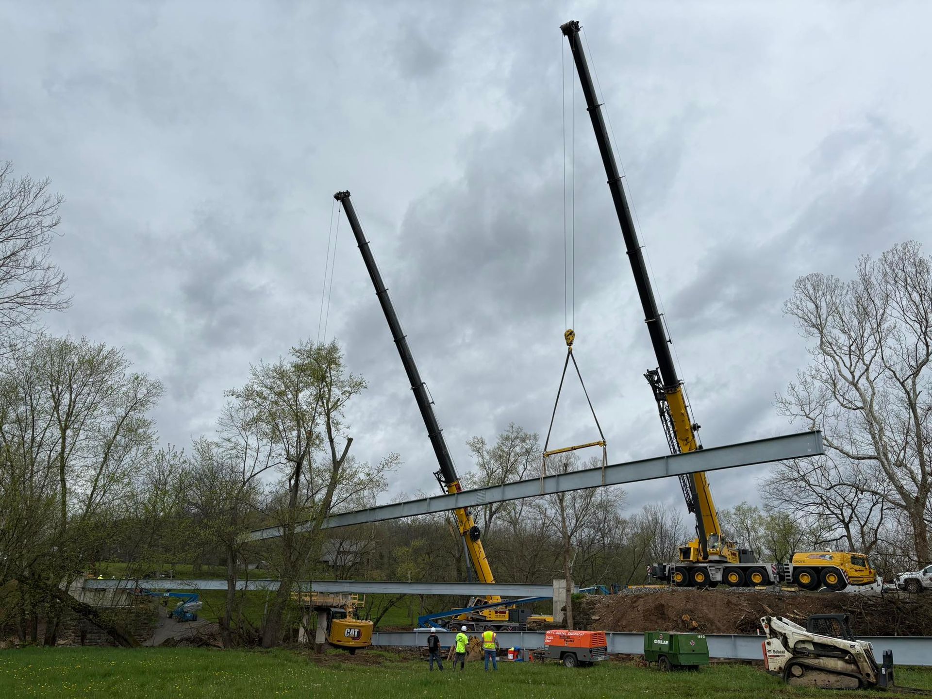 Two cranes lifting a long gray beam at a construction site under cloudy skies