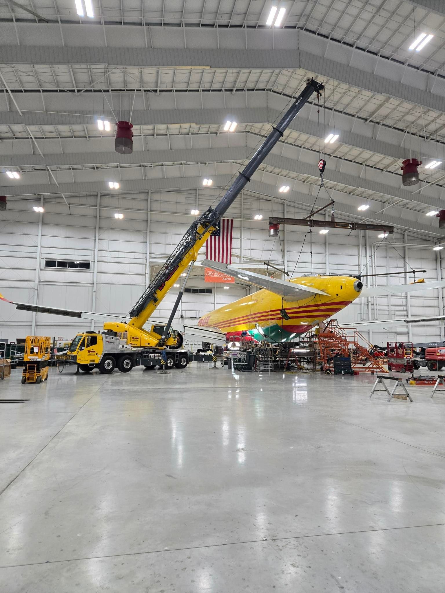 Yellow helicopter being lifted by a crane inside a large hangar with workers nearby