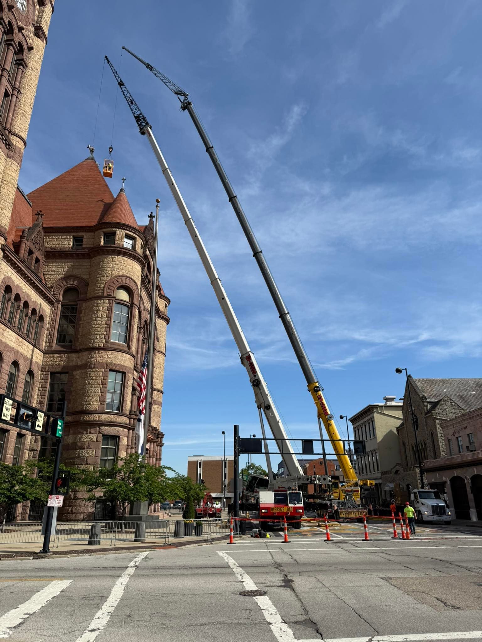 Cranes lifting beams beside a historic stone building on a city street under a blue sky.
