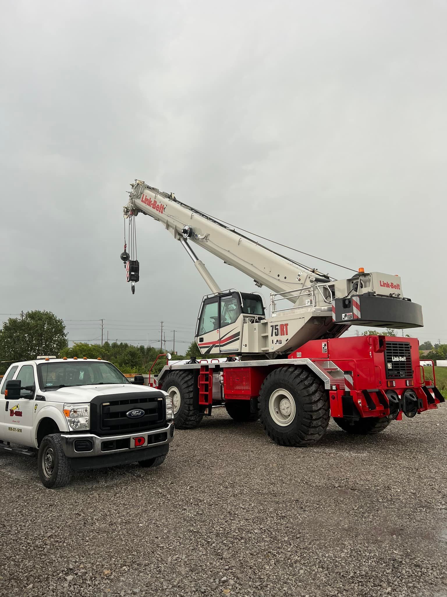 White utility truck and red crane truck parked on gravel under an overcast sky.