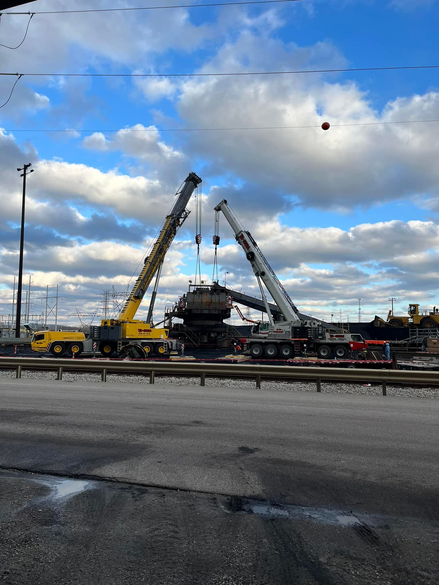 Two cranes lifting a large vehicle at a roadside construction site under a cloudy sky.