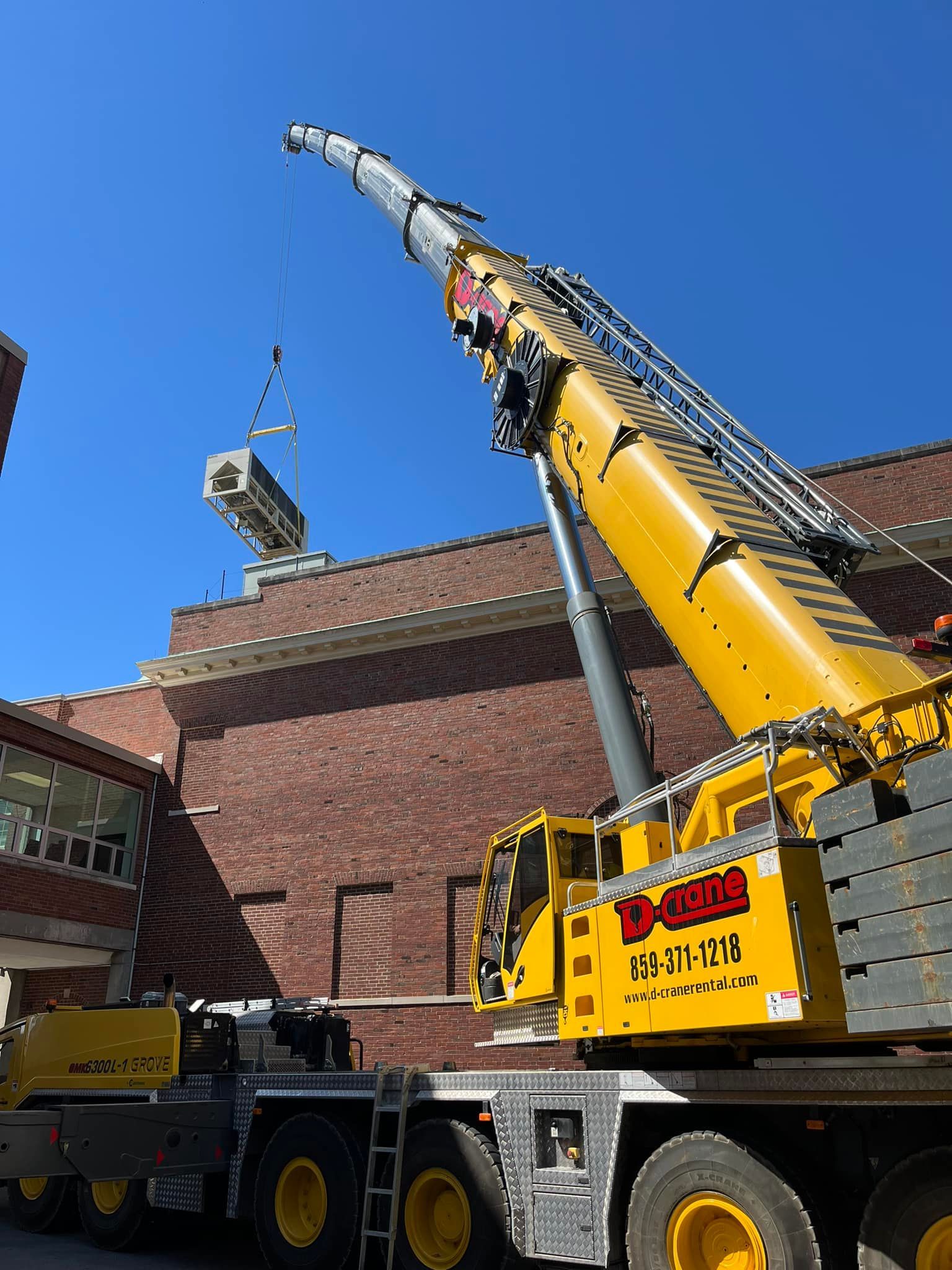 Yellow crane lifting a metal HVAC unit beside a brick building under a blue sky