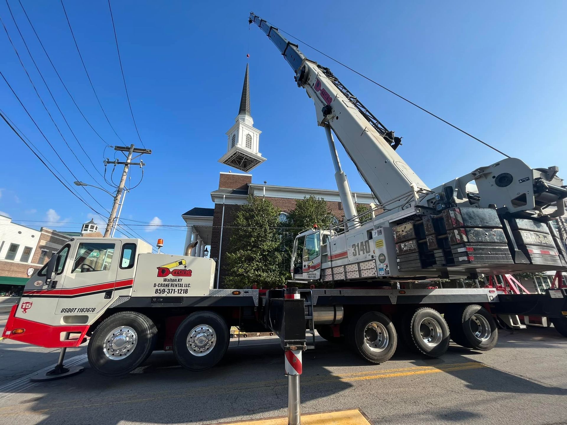 Red and white crane truck parked on a street beside a church tower under a clear blue sky