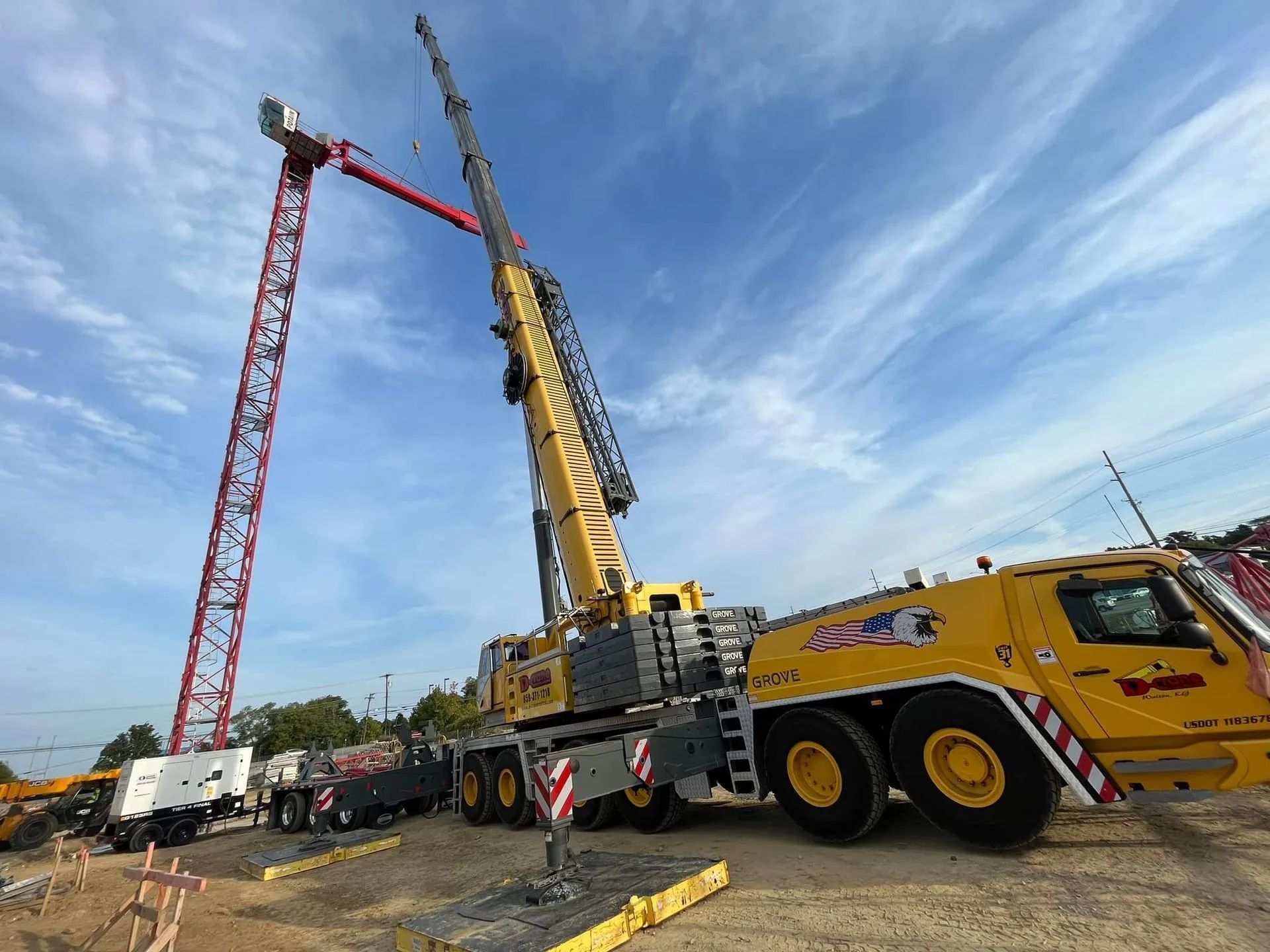 Large yellow crane lifting a red tower crane section at a construction site under a blue sky
