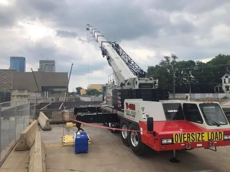 Crane lifting an oversized load on a flatbed truck beside a waterfront construction site