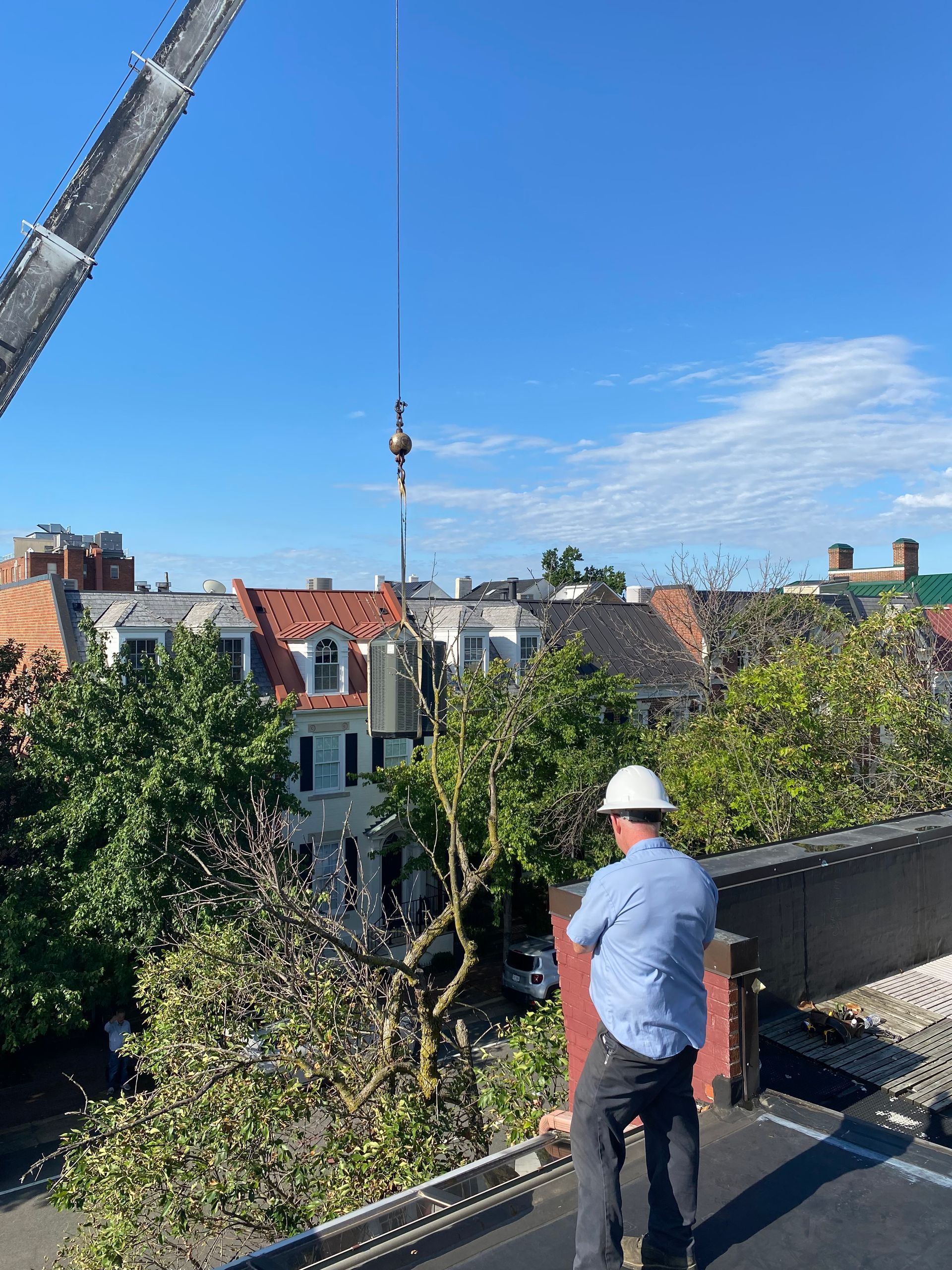 A man is positioned on the rooftop observing a crane hoisting an AC unit