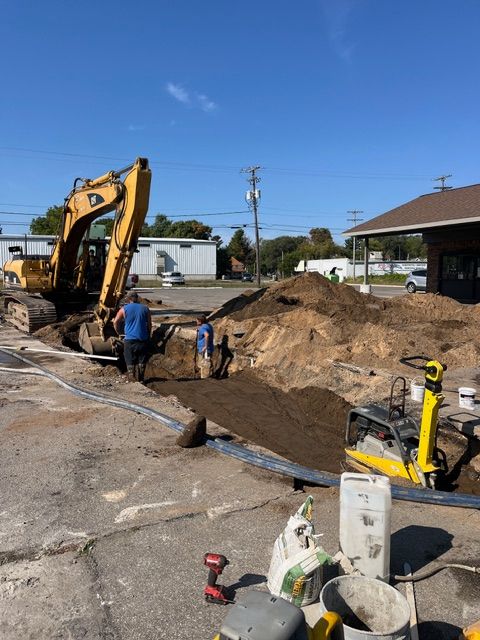 A yellow excavator digs a trench in a parking lot while two workers stand nearby next to a yellow soil compactor.