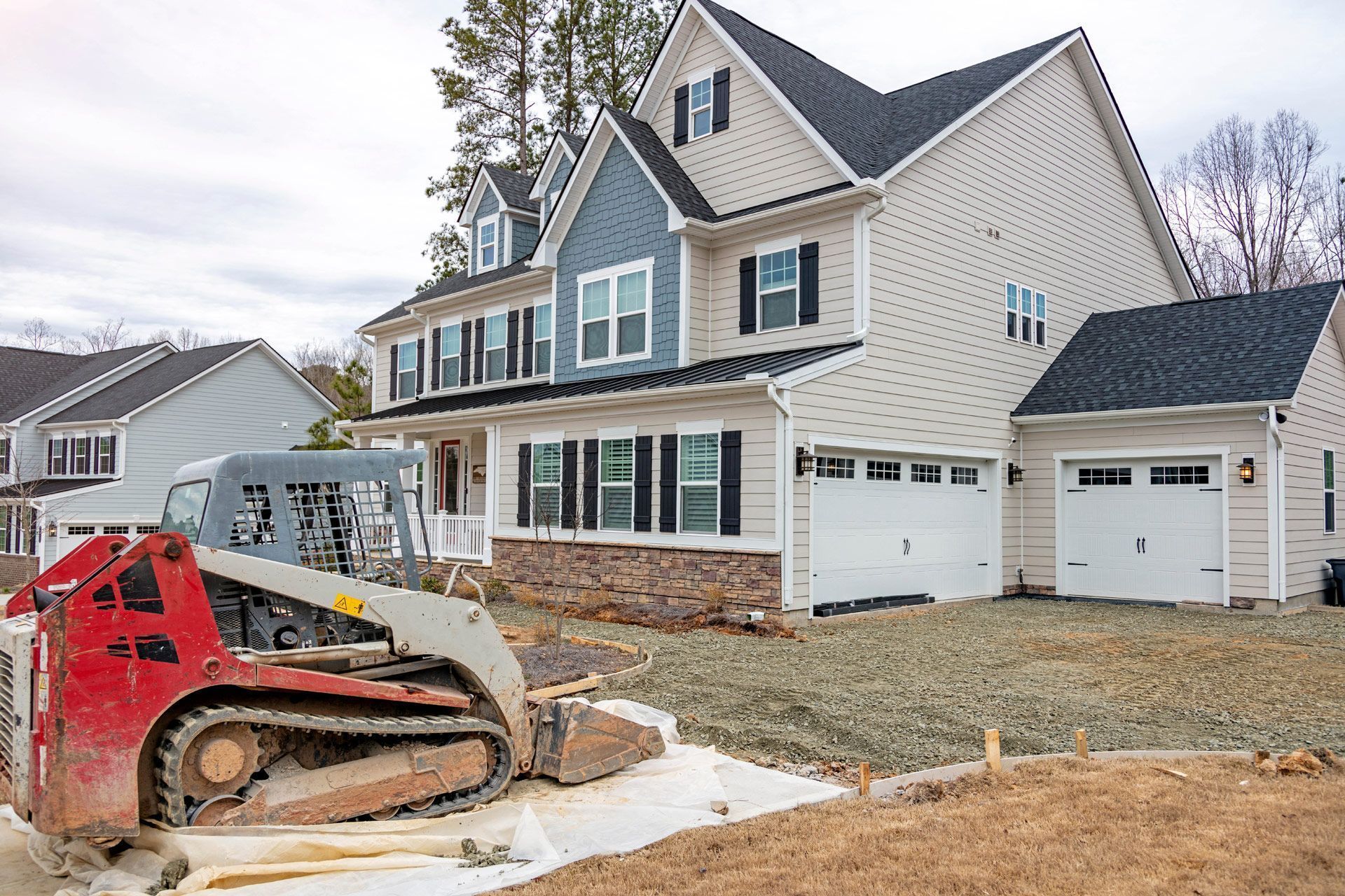 A red and white skid-steer loader sits on a construction site in front of a newly built, two-story suburban house.