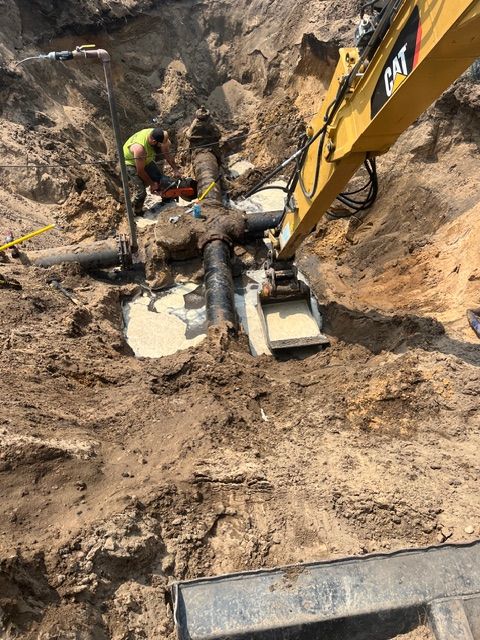 An excavator arm and a worker operate on an exposed underground pipe within a large, sandy construction excavation pit.