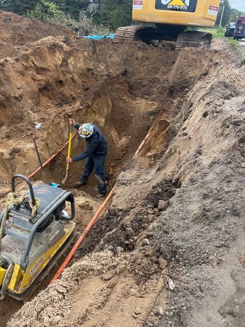 A worker in a hard hat uses a shovel inside a deep, unprotected construction trench next to heavy machinery.