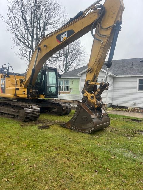 A yellow Caterpillar excavator sits parked on a grassy lawn in front of a white house.