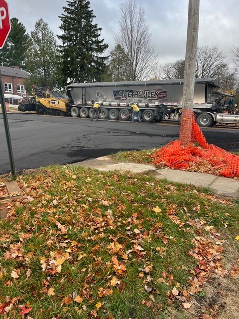 A yellow construction loader next to a parked semi-trailer truck on a paved road with autumn leaves nearby.