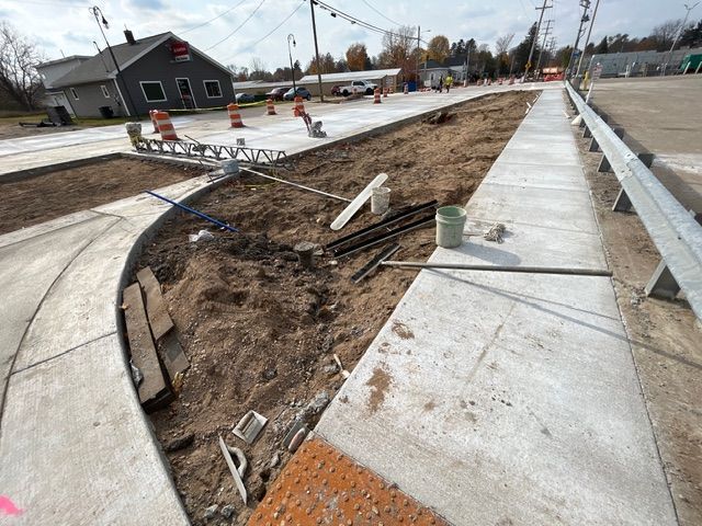 A construction site showing a new concrete sidewalk, a tactile paving ramp, and a dirt-filled island.