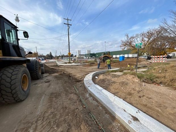 A construction site with a heavy vehicle on the left, new concrete curbing, and workers in safety vests on a sunny day.
