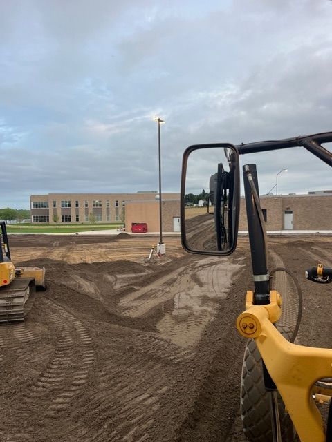 View from the cab of a construction vehicle overlooking a dirt lot with tire tracks and a building in the distance.