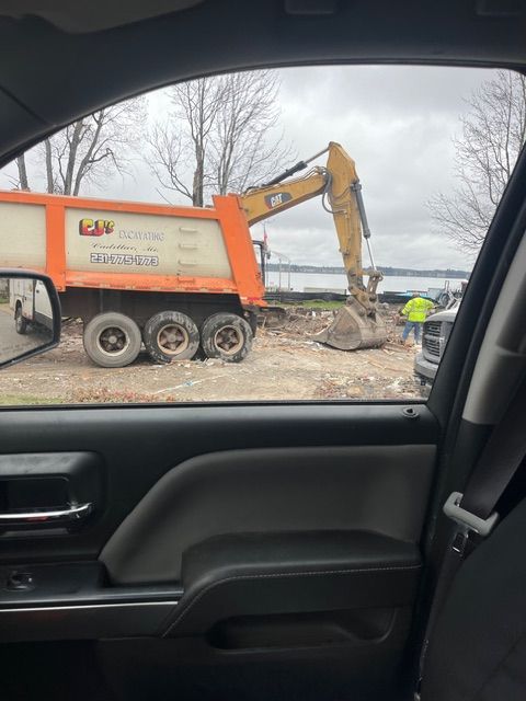 View from a car window showing a yellow CAT excavator loading debris into an orange dump truck at a construction site.