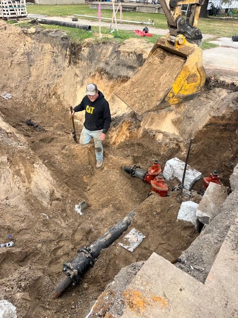 A construction worker stands in a deep trench next to exposed water pipes, with an excavator bucket suspended above.