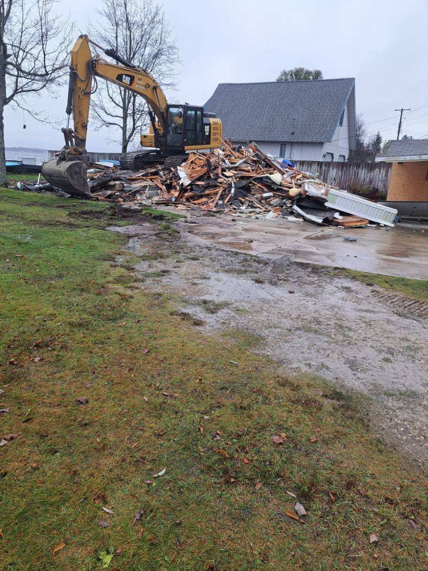 A yellow excavator demolishes a house, leaving a pile of debris on a muddy lot near a grassy area and another building.