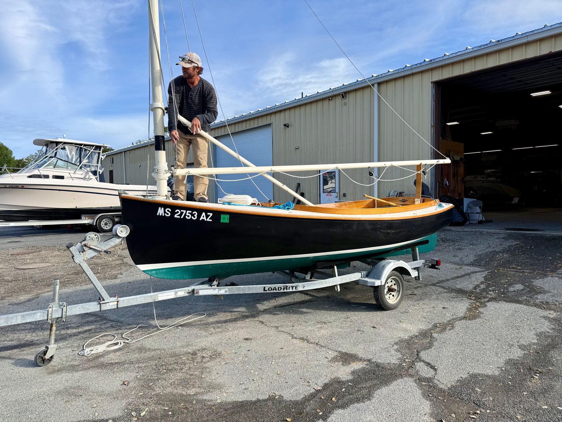 Man on a boat trailer assembling a sailboat. Black hull, green bottom, tan wood deck, building background.