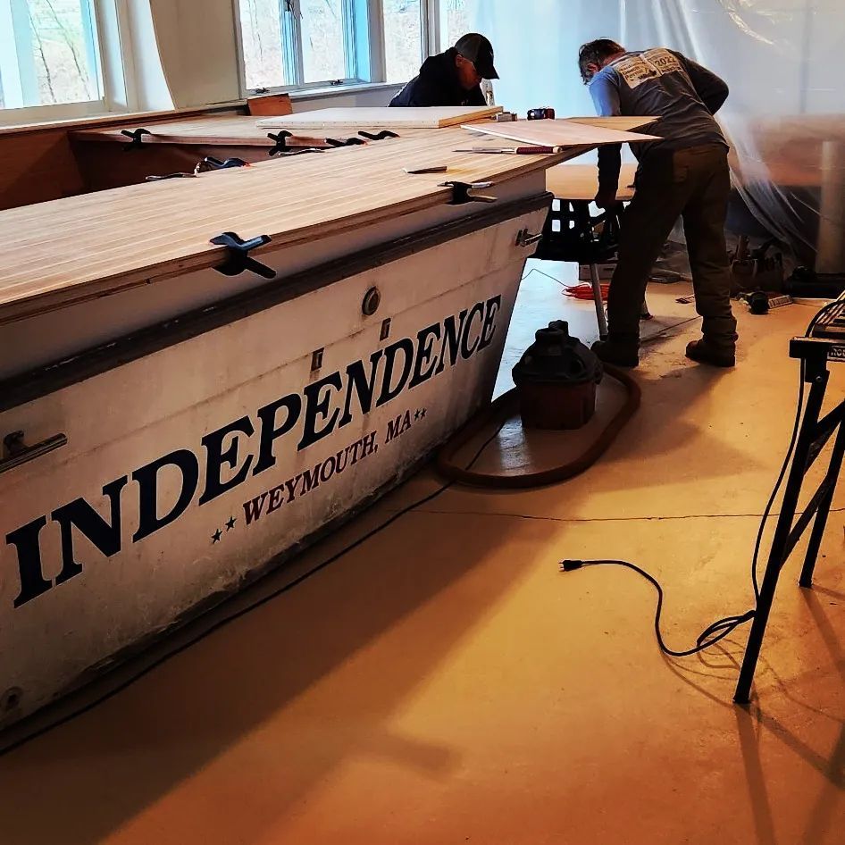 Two people working on a boat named "Independence" indoors. One saws, the other observes.